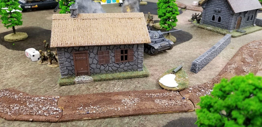 Model of a stone building with a thatched roof, surrounded by miniature tanks and figures on a textured surface.